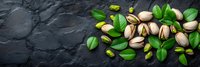 Fresh roasted pistachio nuts with green leaves scattered on dark stone background, top view panoramic banner composition for food photography.