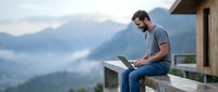 Young bearded man working on laptop outdoors on mountain terrace with scenic valley view. Remote work and digital nomad lifestyle concept.