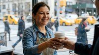Smiling female barista with dark hair and olive complexion hands coffee cup to customer in busy urban cafe with yellow taxis visible through window.