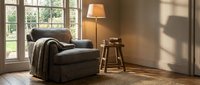 Cozy living room corner with gray upholstered armchair, wooden side table, floor lamp, and natural light from large windows, perfect for interior design and home decor projects.