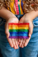 Child's hands holding rainbow pride flag against colorful clothing background, representing support, unity, and acceptance in LGBTQ community movement.