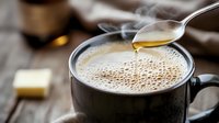 Hot coffee with butter and honey dripping from spoon into black mug on wooden table, steam rising, morning breakfast atmosphere with creamy foam surface.