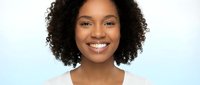 Young African American woman with curly hair smiling warmly at camera wearing white top against light blue gradient background for lifestyle content.