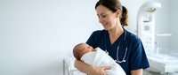 Female healthcare professional in navy scrubs tenderly holding newborn baby in modern hospital room with medical equipment and clean white environment.