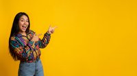 Young woman with long dark hair and South Asian ethnicity wearing colorful patterned shirt and denim, pointing and smiling against bright yellow background for advertising and promotional campaigns.