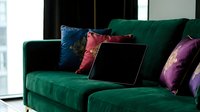 Luxurious emerald green velvet sofa with colorful silk cushions and laptop computer in modern living room interior for home office workspace design.