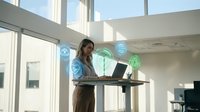 Woman working at standing desk with floating digital network icons representing global connectivity and data transfer in modern office environment.