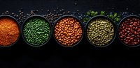 Colorful variety of raw legumes and pulses in black bowls arranged in row on dark background, including red lentils, green peas, kidney beans and chickpeas.