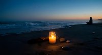 Glowing candle lantern on sandy beach at twilight with ocean waves and silhouetted person creating romantic coastal atmosphere for relaxation and meditation.