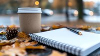 Cozy autumn workspace with takeaway coffee cup, spiral notebook and pencil among fallen maple leaves and pine cones, soft bokeh background creates warm atmosphere.