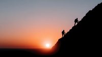 Two hikers climbing steep mountain slope silhouetted against dramatic orange sunset sky. Adventure outdoor recreation concept for travel and fitness content.