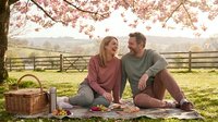 Couple enjoying outdoor picnic under blooming cherry blossom tree in spring countryside setting with basket and food on blanket.