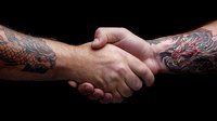 Strong handshake between two tattooed arms with detailed dragon and artistic designs against dark background, symbolizing trust, partnership and agreement.