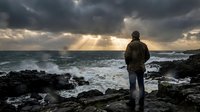 Man standing on rocky coastline gazing at dramatic stormy ocean with golden sunrays breaking through dark clouds, contemplative mood, suitable for motivational and travel content.