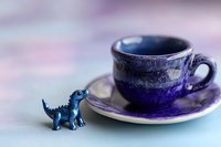 Blue ceramic coffee cup and saucer with small dinosaur figurine on pastel background for morning beverage concepts and playful kitchen decor themes.