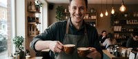 Male barista with dark hair and light skin, wearing a dark apron, smiling while serving two cups of coffee in a modern café with warm lighting, plants, and customers in the background.