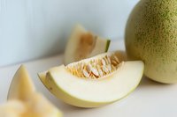 Fresh honeydew melon slice showing juicy flesh and seeds on white background, selective focus on cut piece revealing sweet pale green interior texture.