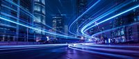 Futuristic city street with glowing blue light trails from traffic creating dynamic motion blur effects through modern urban architecture at night.