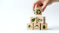 Hand placing wooden cube with recycling symbol on stack of eco-friendly blocks featuring green environmental icons on white background.
