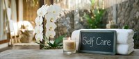 Elegant self care spa setup with white orchids, lit candle, chalkboard sign, and folded towels on wooden surface in serene outdoor setting.