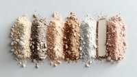Crushed makeup powder swatches in neutral tones from ivory to bronze arranged in horizontal lines on white background, showing texture and pigmentation for cosmetic use.