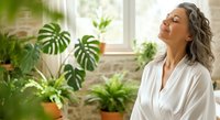 Mature woman with gray hair and light skin tone wearing white shirt surrounded by potted plants near bright window, enjoying peaceful moment in plant-filled home interior.
