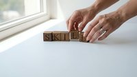 Hands of a middle-aged Caucasian male arranging wooden blocks spelling skills on a white surface near a window, representing professional development and competency building.