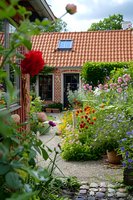 Charming cottage garden with blooming flowers along gravel path leading to brick house with terracotta roof tiles, featuring roses, wildflowers and cottage-style plantings.