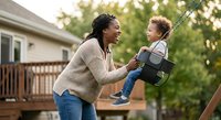 African American mother pushing mixed race toddler son on backyard swing set during golden hour outdoor family playtime activity.