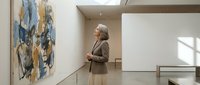 Senior woman with gray hair in brown blazer contemplating abstract artwork in modern gallery space with natural lighting and minimalist design.