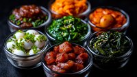 Korean side dishes banchan variety in traditional bowls featuring kimchi, marinated eggs, seasoned vegetables and fermented specialties on dark background.