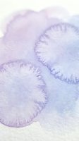 Soft purple watercolor paint spreading on textured paper creating organic flowing shapes with delicate edges and translucent layers for artistic projects.