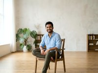 Man with dark hair and beard, appearing to be of Middle Eastern or South Asian descent, wearing a light blue button-up shirt and olive pants, sitting confidently on a wooden chair in a bright, minimalist interior space with potted plants and natural light for corporate portraits or business lifestyle content.