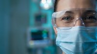 Female healthcare professional wearing protective safety glasses and surgical mask in modern medical facility with blurred equipment background.