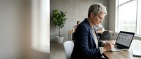 Mature businessman working on laptop in modern office space with natural lighting and plants creating productive work environment.
