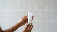 Person with medium skin tone holding white cosmetic tube against white subway tile wall background for skincare product marketing.