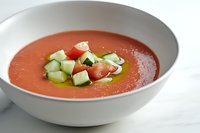 Fresh gazpacho soup garnished with diced cucumber, tomatoes and olive oil in white ceramic bowl on marble surface, cold summer appetizer.