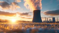 Nuclear power plant cooling towers emitting steam at sunset with frost covered grass in foreground, dramatic orange sky and sunburst through winter trees.