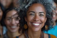 Confident mature African American woman with curly gray hair smiling brightly, wearing ethnic jewelry against blurred background of diverse people.