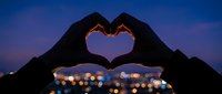 Hands forming heart shape silhouette against twilight sky with city lights bokeh background. Romantic gesture concept for love and affection themes.