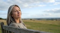 Mature Caucasian woman with gray hair sitting on bench outdoors, gazing thoughtfully at horizon with serene expression in natural countryside.