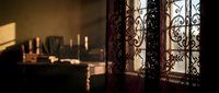 Warm sunlight streaming through ornate wrought iron window grille casting decorative shadows in cozy interior room with candles and vintage atmosphere.