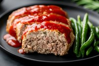 Sliced homemade meatloaf topped with rich tomato sauce served on black plate with fresh steamed green beans, comfort food dinner photography.