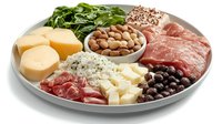 Gourmet appetizer platter with assorted meats, cheeses, nuts, olives and fresh spinach leaves arranged on round ceramic plate, Mediterranean style charcuterie board.