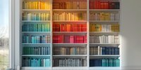 Colorful rainbow bookshelf arrangement with books organized by color spectrum from yellow to blue, creating striking visual display against white wall and window.
