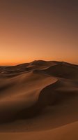 Vast desert sand dunes glow under a warm orange sunset sky, casting deep shadows across the rolling landscape.