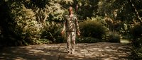 Young Caucasian man walking on sunny garden path surrounded by lush green foliage and dappled sunlight creating peaceful outdoor atmosphere for wellness content.