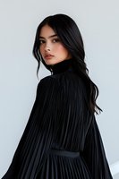 Asian woman with long dark hair wearing black pleated dress looking over shoulder against light background. Elegant fashion portrait with minimalist styling.