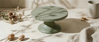 Green marble cake stand on white fabric with dried flowers and ceramic mug creating elegant minimalist food photography setup for bakery displays.