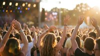 Crowd of people with raised hands enjoying outdoor music festival at sunset with stage lights and warm golden hour atmosphere.